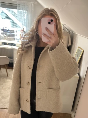 Beige kappa - Säljer en beige kappa med krage och stora knappar framtill. Kappan har två rejäla fickor och är gjord i mjukt, fluffigt material som ger en cozy vibe. Perfekt för kyliga dagar när du vill vara både varm och stylish. 