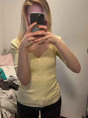Ljusgul ribbad topp med v-ringning - Säljer en ljusgul ribbad topp med v-ringning och korta ärmar. Perfekt för dig som gillar en enkel och stilren look. Toppen har en slim passform och är gjord i mjukt bomullsmaterial som känns skönt mot huden.