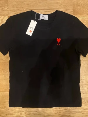 Svart t-shirt från Ami Paris - Snygg svart t-shirt från Ami Paris med röd broderad logga i form av ett hjärta och bokstaven A på bröstet. Klassisk rund hals och korta ärmar. Tillverkad i mjuk bomull för en skön känsla och stilren look.