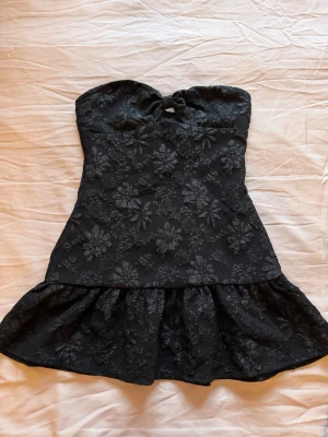 Svart axelbandslös klänning med blommönster - Säljer en svart axelbandslös klänning med volangkjol och broderat blommönster. Klänningen har en liten knutdetalj vid bysten och är tillverkad i ett tyg med struktur. Perfekt för dig som gillar en feminin och trendig look.