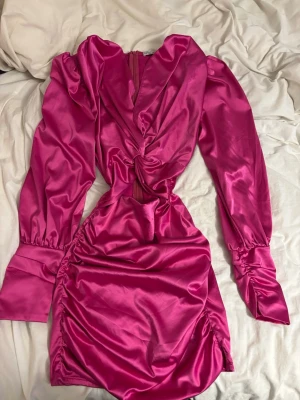 Rosa satin klänning från Glamaker - Snygg kort rosa klänning i satin från Glamaker med långa puffärmar, knytdetalj framtill och lätt drapering i midjan. Klänningen har en v-ringning och markerade manschetter för en trendig look.Liten i storlek