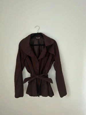 Brun trenchcoat -  brun trenchcoat med bred krage, dubbelknäppning och tillhörande knytskärp i midjan. 
