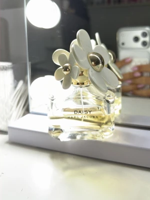Daisy parfym från Marc Jacobs - Daisy från Marc Jacobs är en ikonisk parfym i en genomskinlig glasflaska med guldfärgad detalj och vita blommor på locket. Doften är fräsch och feminin, perfekt för dig som gillar eleganta och lekfulla parfymer. Flaskan är snygg att ha framme på sminkbordet.