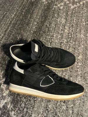  Philippe Model trainers - Stilrena svarta sneakers från Philippe Model med detaljer i vitt och grått. Ovandelen är i mocka och textil, med en ikonisk sköldlogga på sidan. Yttersulan är vit med beige inslag och skon har svarta skosnören. Perfekta för dig som gillar en clean och sportig look. Skriv vid funderingar och pris är förhandlingsbart vid snabb affär