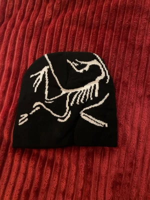 Svart arcteryx mössa - Cool svart mössa med vit stickad fossil- eller skelettdesign på framsidan. Mössan är mjuk och stretchig, perfekt för att hålla värmen och ge en unik stil till din outfit. Passar dig som gillar streetwear och vill sticka ut lite extra.
