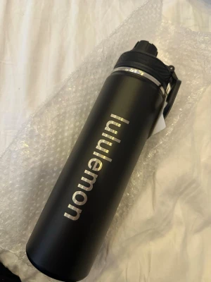 Svart vattenflaska från Lululemon - Snygg svart vattenflaska från Lululemon i rostfritt stål med silvrig logga längs sidan. Flaskan har ett praktiskt lock med bärhandtag och pip för enkel användning. Perfekt att ha med till gymmet, skolan eller på språng.