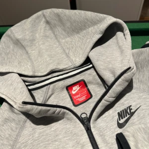Grått Nike Tech juniorstorlek L/XL  - Snygg grå zip-hoodie från Nike med svart logga på bröstet i storlek L ca 147-158 cm. Byxan är i juniorstorlek XL, ca 155-165 cm. Byxorna är slitna på knäna, men stället ser fortfarande fräscht ut. 