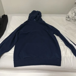 Mörkblå hoodie med vit rand - Mörkblå hoodie med huva och en vit rand längs ena ärmen. Klassisk känguruficka framtill och ribbade muddar vid ärmslut och nederkant. Perfekt för dig som gillar en sportig och avslappnad stil.