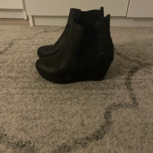 Svarta kilklacksboots - Säljer ett par svarta boots med kilklack och rund tå. Skorna har en stilren design med resår på sidorna och är tillverkade i ett material som liknar skinn. Perfekta för dig som vill ha lite extra höjd och en klassisk look.