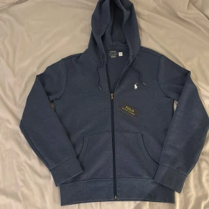 Blå hoodie från Polo Ralph Lauren - Säljer en snygg marinblå zip hoodie från Polo Ralph Lauren. Perfekt för chill dagar eller när du vill ha en stilren look. NYPRIS 2 395kr! Man får med Ralph lauren kasse.