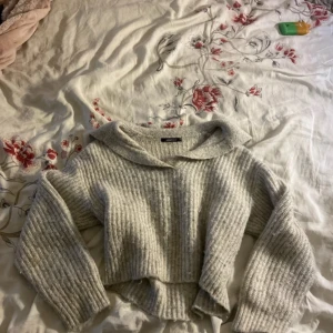 Ljusgrå stickad tröja från Gina Tricot - Supermysig ljusgrå stickad tröja från Gina Tricot i ribbat material. Tröjan har en bred krage, croppad passform och långa ärmar. Perfekt att styla med jeans eller kjol för en chill look.