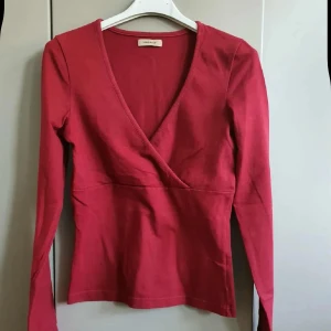 Röd omlottopp från Vero Moda - Snygg röd omlottopp från Vero Moda med lång ärm och djup v-ringning. Toppen är i mjuk bomull och har en figurnära passform som framhäver siluetten. Perfekt att styla med jeans eller kjol för en trendig look.