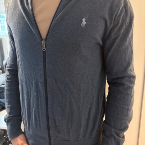 Blå kofta från Polo Ralph Lauren - Snygg marinblå kofta från Polo Ralph Lauren med dragkedja framtill och broderad logga på bröstet. Tröjan är långärmad och har en klassisk, normal passform. Perfekt att slänga på sig när det är lite kyligt ute.