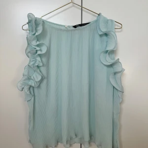 Ljusblå volangblus från Zara - Supersöt ljusblå blus från Zara med plisserat tyg och volangdetaljer längs axlarna. Blusen är ärmlös och har en rund halsringning med knapp i nacken. Perfekt för dig som gillar romantiska och luftiga plagg.