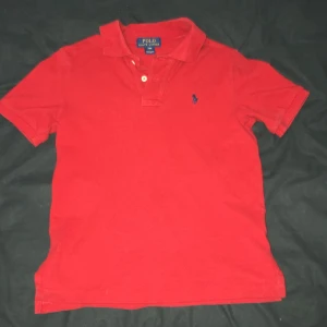 Röd pikétröja från Polo Ralph Lauren - Snygg röd pikétröja från Polo Ralph Lauren med klassisk krage och två knappar vid halsen. Tröjan har korta ärmar och den ikoniska blå logon broderad på bröstet. Perfekt för dig som gillar stilren och sportig look.