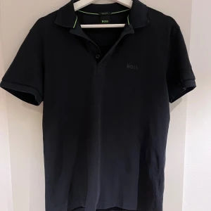 Svart pikétröja från BOSS, storlek S - Svart pikétröja från BOSS i storlek S. Klassisk krage med två knappar och diskret BOSS-logga på bröstet. Pikén har gröna detaljer på insidan av kragen och är tillverkad i mjuk bomull. Passformen är normal. Inga fläckar eller tecken på användning!