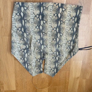 Snakeprint korsettopp från Pimkie - Säljer en snygg korsettopp från Pimkie med beige och svart snakeprint. Toppen har en unik form med spetsig nederkant och metallring i halsen som ger en cool detalj. Den är ärmlös och har knytning i nacken. Perfekt för dig som vill sticka ut.