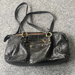 Svart axelväska i krokopräglat skinn - Cool svart axelväska från The Monte i krokopräglat skinn med guldiga metalldetaljer. Väskan har dubbla axelband, dragkedja upptill och flera fack med dragkedja både fram och bak. Perfekt storlek för att få plats med allt du behöver. Lite fläckar inuti annars fint skick