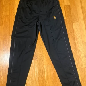 Svarta träningsbyxor från Nike  - Svarta träningsbyxor från Nike med broderad logga i rött och gult på låret. elastisk midja och diskreta sömdetaljer längs benen. Perfekta för sport eller chill. 