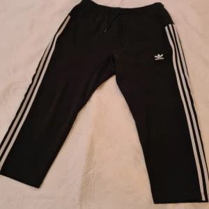 Svarta Adidas mjukisbyxor med vita ränder - Svarta mjukisbyxor från Adidas med klassiska vita ränder längs sidorna och Adidas-logga på låret. Byxorna har snörning i midjan och praktiska fickor med dragkedja. Tillverkade i Primeblue-material med återvunnen plast från havet.