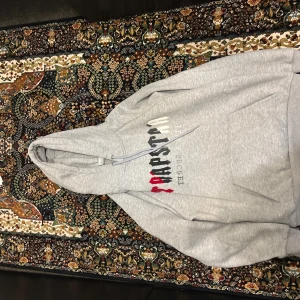 Grå hoodie från Trapstar i storlek L - Säljer en grå hoodie från Trapstar med tryck i svart, vitt och rött framtill. Tröjan har huva med snörning och en stor magficka. Perfekt för dig som gillar streetwear och vill ha en bekväm och snygg look.