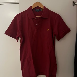 Vinröd pikétröja från Ralph Lauren - Säljer en vinröd pikétröja från Polo Ralph Lauren i modellen Custom Slim Fit. Tröjan har korta ärmar, klassisk krage och två knappar framtill. Ikoniska gula loggan broderad på bröstet. Perfekt för dig som gillar stilren och sportig look.