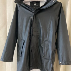 Svart regnjacka från Cross Sportswear - Svart regnjacka från Cross Sportswear med huva och tryckknappar framtill. Jackan har raka fickor och logga på ärmen. Materialet är syntetiskt och jackan är helsvart med en stilren look, perfekt för regniga dagar.