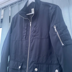 Vindjacka från Jack & Jones - Snygg vindjacka från Jack & Jones Premium med dragkedja framtill, fickor med tryckknappar och en extra ficka med dragkedja på ärmen. Jackan har ståkrage och raka ärmar, perfekt för dig som gillar stilren och enkel design | Passar för någon runt 175-182 | Tveka it fråga om du har funderingar 👍