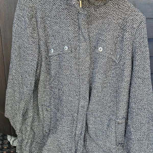 Gråmelerad rock med knappar - Snygg gråmelerad rock med klassiskt rutigt mönster och knappar framtill. Jackan har två bröstfickor med lock och knappar samt två sidofickor. Perfekt för dig som gillar en stilren och tidlös look.
