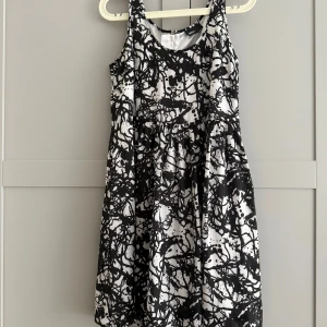 Svartvit kort klänning från Zoul - Cool svartvit klänning från Zoul med abstrakt mönster och rund halsringning. Modellen är ärmlös och har en markerad midja samt dragkedja i ryggen. Materialet är glansigt och känns lyxigt, perfekt för dig som gillar att sticka ut.