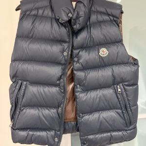 Mörkblå dunväst från Moncler - Snygg mörkblå dunväst från Moncler med quiltad design och hög krage. Västen har dragkedja och tryckknappar framtill samt två fickor med dragkedja. Klassisk Moncler-logga på bröstet och glansig finish. Perfekt för lager-på-lager-stil.
