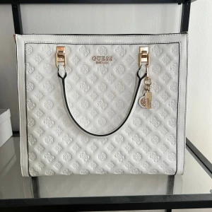 Vit handväska från Guess med mönster - Snygg vit handväska från Guess med präglat logomönster och guldfärgade detaljer. Väskan har svarta kanter, avtagbar axelrem och dragkedja upptill. Insidan är fodrad med beige tyg och Guess-loggor. Perfekt för dig som vill ha en stilren och trendig väska.