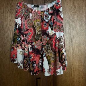 Färgglad mönstrad kjol från Ecaline - Säljer en kort kjol från Ecaline med färgstarkt paisleymönster i rött, grönt, vitt och mörkt rött. Kjolen har en lätt utsvängd form och är tillverkad i mjuk polyester. Perfekt för dig som gillar unika och bohemiska plagg med mycket färg och detaljer.