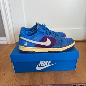 Nike Dunk Low SP Undefeated Blue/Plum - Nike Dunk Low SP Undefeated i blå mocka och skinn med lila paneler och vit Swoosh. Snygga detaljer med Undefeated-logga på hälen, gul vintage-inspirerad sula och 2 extra skosnören följer med. Riktigt feta skor, skick 9/10. Om du har fler frågor eller är intresserad, skriv till mig privat