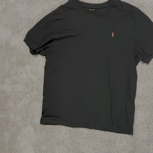 Svart t-shirt från Polo Ralph Lauren - Svart t-shirt från Polo Ralph Lauren med klassisk röd broderad logga på bröstet. T-shirten har rund halsringning och är tillverkad i mjuk bomull. Enkel och stilren modell som passar perfekt till jeans eller shorts.