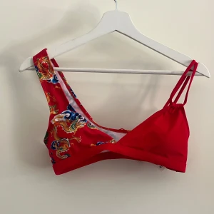 Röd bikiniöverdel med drakmönster - Röd bikiniöverdel från SHEIN med drakmönster i gult, blått och grönt. Ena axelbandet är dubbelt och den andra är bredare.