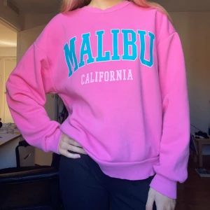 Rosa sweatshirt med tryck🩷 - Cool rosa sweatshirt med stort turkost och vitt 'MALIBU CALIFORNIA'-tryck på bröstet. Oversized fit med ribbade muddar och rund hals. Perfekt för dig som gillar rosa🩷🤩Mjuk och skön! (Har en liten fläck vid armen men är knappt märkbart) Orginalpris 249kr men säljer för 149kr