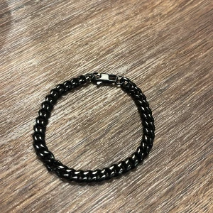 Svart kedjearmband i metall - Stilrent svart armband med kedjelänkar i metall. Armbandet har en klassisk karbinhake som lås och en glansig finish som ger en cool vibe. Perfekt accessoar för att lyfta din outfit med en edgy touch. Den rostar inte