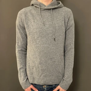 Grå stickad hoodie med huva - Snygg grå stickad hoodie med huva och snörning. Tröjan har långärmade raglanärmar och ribbade muddar vid ärmslut och nederkant. Perfekt för dig som gillar en chill och enkel stil. Passar dig som vill ha en mjuk och varm tröja med relaxed vibe.