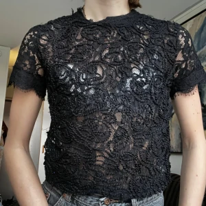 Svart spetsblus från Zara - Supersnygg svart blus från Zara med genombruten spets och korta ärmar. Blusen har rund hals, dragkedja i nacken och fina broderade detaljer. Perfekt att styla med jeans eller kjol för en trendig look.