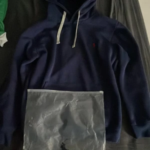 Marinblå hoodie från Ralph Lauren - Säljer en marinblå hoodie från Ralph Lauren med klassisk huva och vita snören. Tröjan har en liten röd broderad logga på bröstet och en stor ficka framtill. Perfekt för dig som gillar stilren och sportig look.