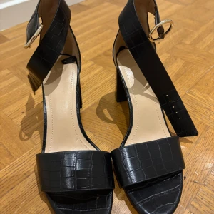 Svarta klackade sandaler med crocomönster - Stilrena svarta sandaler med bred rem över tårna och justerbart ankelband med guldfärgat spänne. Skorna har crocomönstrad yta och blockklack som ger extra höjd. Perfekta för dig som vill ha en trendig och elegant look.