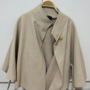 Beige kappa från Missay med bälte - Stilren beige kappa från Missay med bred krage och knäppning med både vit och guldig knapp. Jackan har ett bälte i midjan och vida ärmar. Onesize.