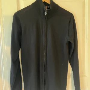 Svart kofta från Oscar Jacobson - Svart, stilren kofta från Oscar Jacobson med hel dragkedja och ribbad krage. Tillverkad i Italien i mjukt stickat material. Sitter snyggt med normal passform och långa ärmar. Perfekt till jeans eller chinos för en clean look. Den har ett mindre märke som man kan se på bild 3 och därför billigare. Köpt för runt 2000kr på deras hemsida. 