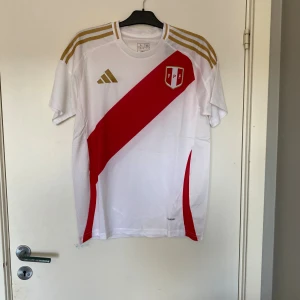 Peru landslagströja Adidas L - Snygg Peru landslagströja från Adidas i storlek L. Tröjan är vit med en röd diagonal rand och guldiga detaljer på axlarna. Klassisk rund hals och korta ärmar. Tillverkad i lätt och ventilerande polyester, perfekt för match eller träning.