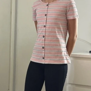 Randig rosa blus med knappar - Säljer en söt randig blus i ljusrosa med vita och mörka ränder. Blusen har korta ärmar, rund hals och knappar hela vägen framtill. Materialet är ribbat och känns mjukt mot huden. Perfekt för dig som gillar en chill och fräsch stil.