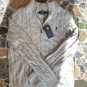 Grå kabelstickad tröja Polo Ralph Lauren - Kabelstickad grå tröja från Polo Ralph Lauren med halv dragkedja och hög krage. Klassisk design med broderad logga på bröstet och ribbade muddar. Perfekt för lager-på-lager och snygg till jeans eller chinos.