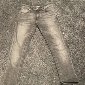 Grå skinny jeans från Nudie Jeans - Snygga grå jeans från Nudie Jeans i slim fit. Jeansen har klassisk femficksdesign, svarta vågdetaljer på bakfickorna och en tvättad look. Perfekta för dig som gillar en smal siluett och vill ha ett par stilrena jeans i garderoben. Storlek L30 W25 men de har krympt i tvätten så skulle säga mer L29-L28