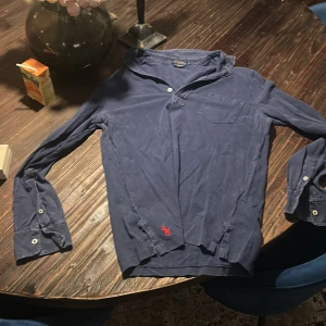 Blå pikétröja från Polo Ralph Lauren - Snygg marinblå långärmad pikétröja från Polo Ralph Lauren i lätt bomullsmaterial. Klassisk krage, knappslå och bröstficka. Ikoniska röda loggan broderad nertill. Perfekt för en chill och stilren look.