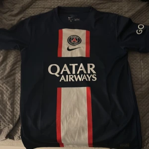 PSG matchtröja Nike Dri-Fit L - Paris Saint-Germain fotbollströja från Nike i storlek L. Tröjan är marinblå med vit och röd vertikal rand på framsidan, PSG-logga och Qatar Airways-tryck. Kortärmad och tillverkad i Dri-Fit material som andas. Snygg detalj med 'GOAT' på ärmen.